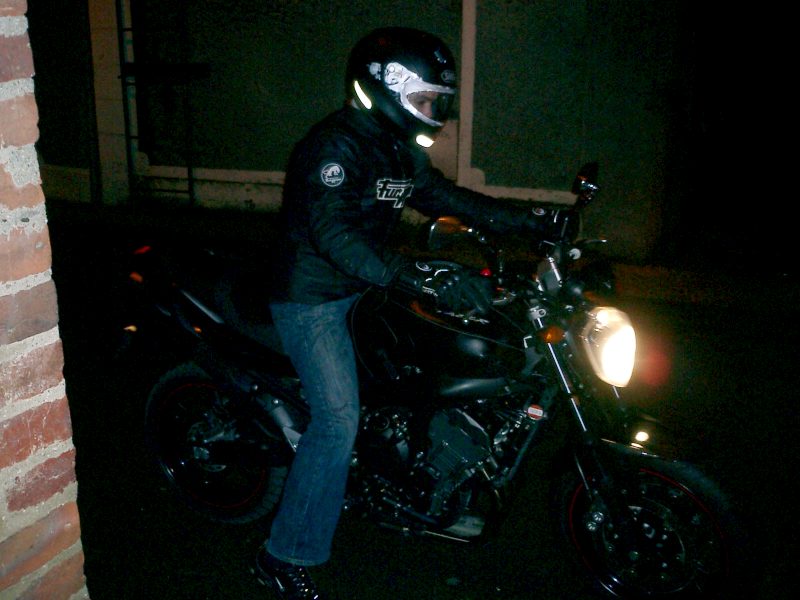 Le pilote casqué, blouson fermé, enfourche la moto phare allumé, de nuit.